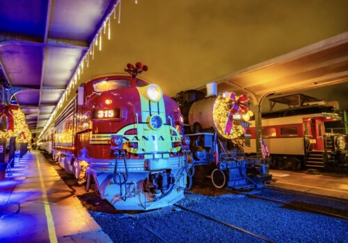 The Santa Fe 315 diesel locomotive decorated with Christmas wreaths and string lights stands on the indoor platform at night, with a steam locomotive and additional holiday-decorated rail cars visible alongside it.