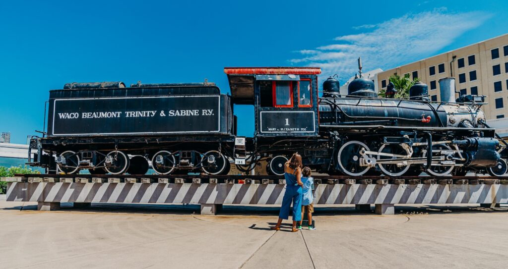 A woman and child view a historic black steam locomotive outside the Galveston Railroad Museum.