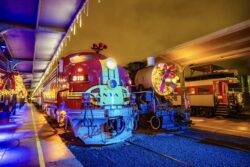 The Santa Fe 315 diesel locomotive decorated with Christmas wreaths and string lights stands on the indoor platform at night, with a steam locomotive and additional holiday-decorated rail cars visible alongside it.