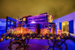 A historic Waco Beaumont Trinity & Sabine Railway locomotive and an antique wagon decorated with glowing gift boxes, garland, and string lights in the museum courtyard at night, bathed in purple and gold light.