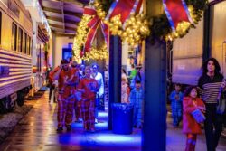 Families in pajamas walk along the museum's indoor train platform beneath decorated wreaths with red bows and string lights, with vintage passenger cars on both sides.