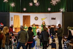 Three performers in matching green dresses sing at vintage microphones on a decorated stage while families and children watch from the floor of the museum's main hall, which is hung with starburst light fixtures.