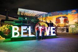 Two children in pajamas stand inside a lit circular arch within a large illuminated BELIEVE sign, with a Case steam tractor and a Santa Fe mural visible in the background.
