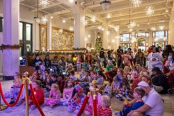 A large crowd of children and adults in pajamas fills the museum's historic depot hall, seated on the floor and standing along the walls, watching a performance beneath starburst light fixtures and holiday decorations.