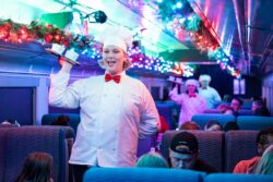 A performer dressed as a chef in a white uniform and red bow tie carries a tray of hot chocolate cups down the aisle of a decorated passenger car, with a second chef visible in the background beneath garland and colored lights.