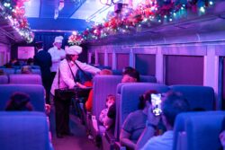 A chef performer leans across a seat row to serve guests inside a holiday-decorated passenger car, with a conductor and second chef visible in the background and colorful garland lining the overhead rails.