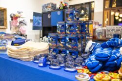A blue-draped merchandise table displaying stacked Polar Express ornament boxes, snow globes, conductor hats, mittens, and collectible coins inside the museum depot.