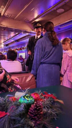 Video of the conductor in uniform leans toward a young guest seated at a table inside a decorated passenger car, with garland, holiday lights, and other guests visible throughout the car.