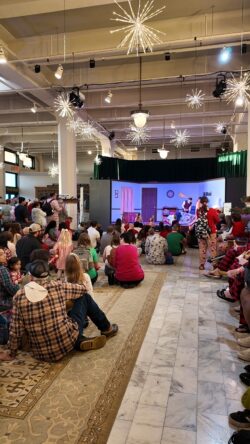 Video of a large crowd of children and adults in pajamas fills the museum's historic depot hall, seated on the floor and standing along the walls, watching a performance beneath starburst light fixtures and holiday decorations.