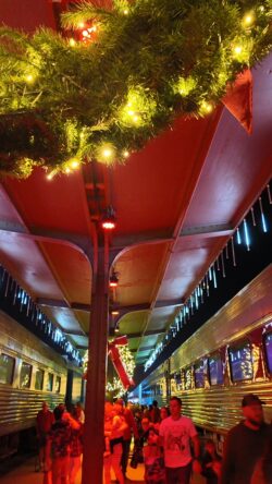 Video of many Families in pajamas walk along the museum's indoor train platform beneath decorated wreaths with red bows and string lights, with vintage passenger cars on both sides.