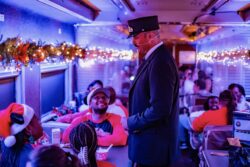 A conductor in uniform leans toward a young guest seated at a table inside a decorated passenger car, with garland, holiday lights, and other guests visible throughout the car.